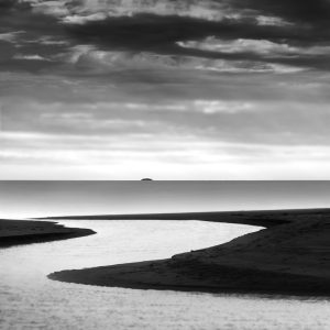 A stream meandering into the sea with an island in the distance under a swirling cloud