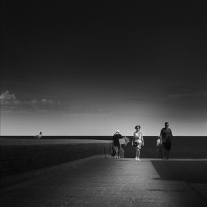 Four people walking on a stone-paved street and a pigeon on a wall with sea and sky as background