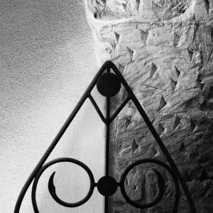 Triangular wrought-iron chairback against a white wall that has irregular etchings on its right half