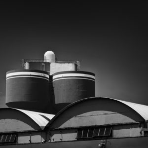 Two dark cylindrical tanks above the two white curved roofs of a factory