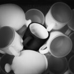 Dark mug with white interior at centre of heap of white mugs and piggy banks.