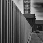 Squared-shaped skyscraper surrounded by clouds in background, tall metal-bar fence stretching in perspective from left side.