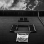 View from the ground of tricolour flag hanging from an open window; another closed window, eaves and clouds are above it.
