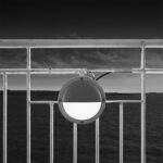Circular lamp with dark top half attached to the white railing of a ferry, with sea and sky in background.
