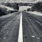 White road line on straight two-lane road leading to hills under cloudy sky.