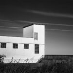 White pumping station with graffiti in lower left corner and four glass-block windows; grass in foreground and striped clouds.