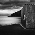 Wooden shack with slanting walls on beack, with sea, promontory and horizon in background.