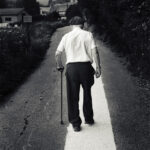 Old man wearing shite shirt and dark coppola and pants seen from behind walking on a large white stripe with stick in left hand
