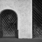 White wall with wooden arched door on the left, large wooden door filling right side of picture.