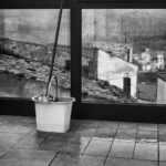 Mop and bucket set leaning against window facing roofs on rainy day.