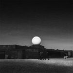 Large, white dome on top of North Cape Hall - a dark, low stone building at North Cape, Norway. Three men in dark motorcycle suits walk along a wall under the dome and a group with a white dog stands on the left.
