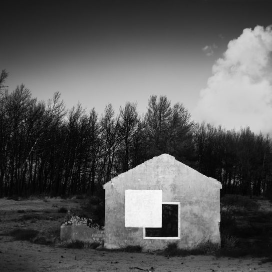 A black and a white square painted on the wall of a ruined house with dead trees and clouds in the background