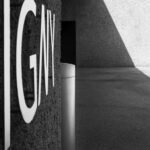 Large clear I-G-N-Y metal letters on wall on left; a metal bin in the centre is aligned with a dark door in a wall